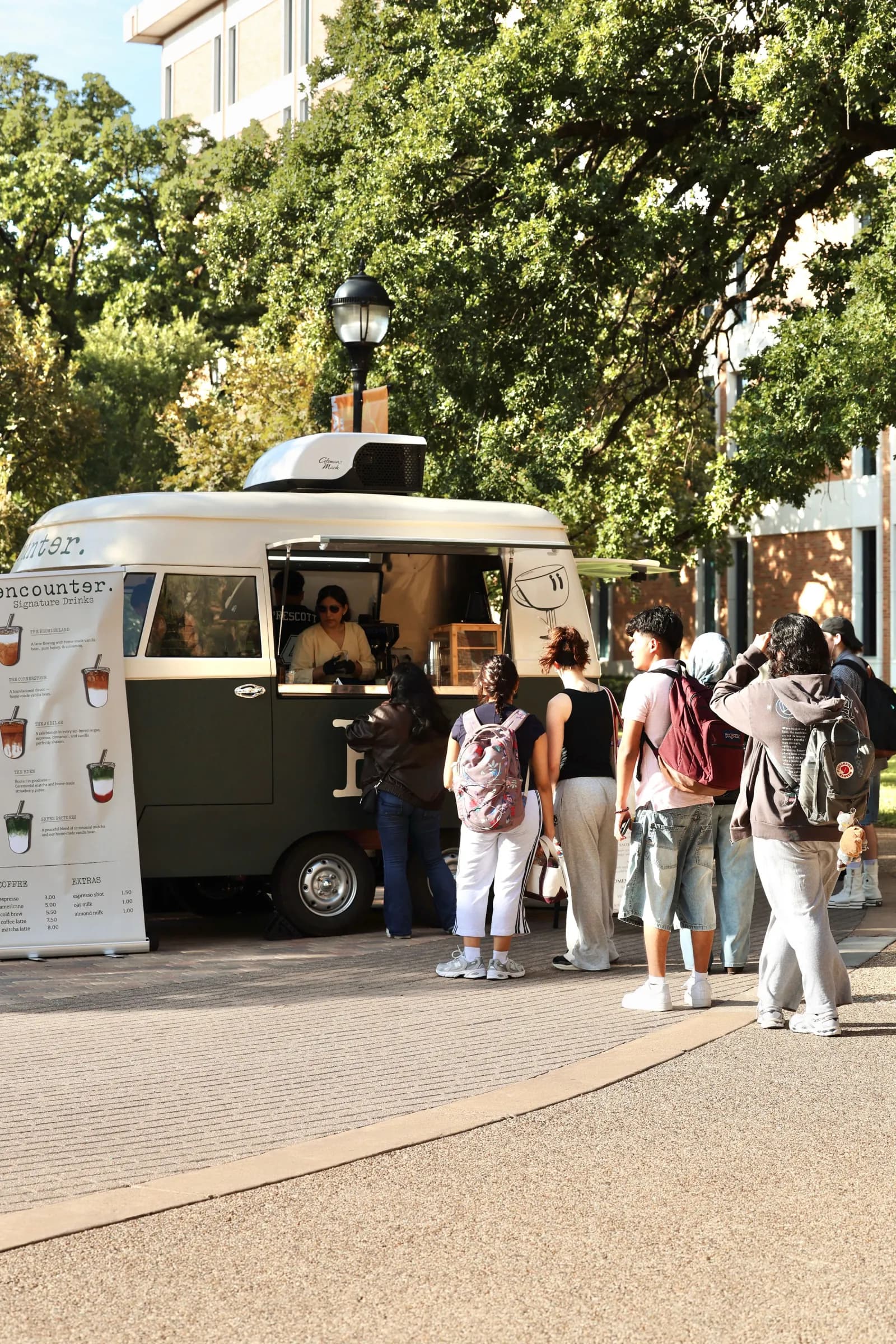 Guests lined up at the Encounter Coffee vintage VW bus coffee trailer, with the signature drinks menu beside the serving window, at an outdoor Dallas–Fort Worth event