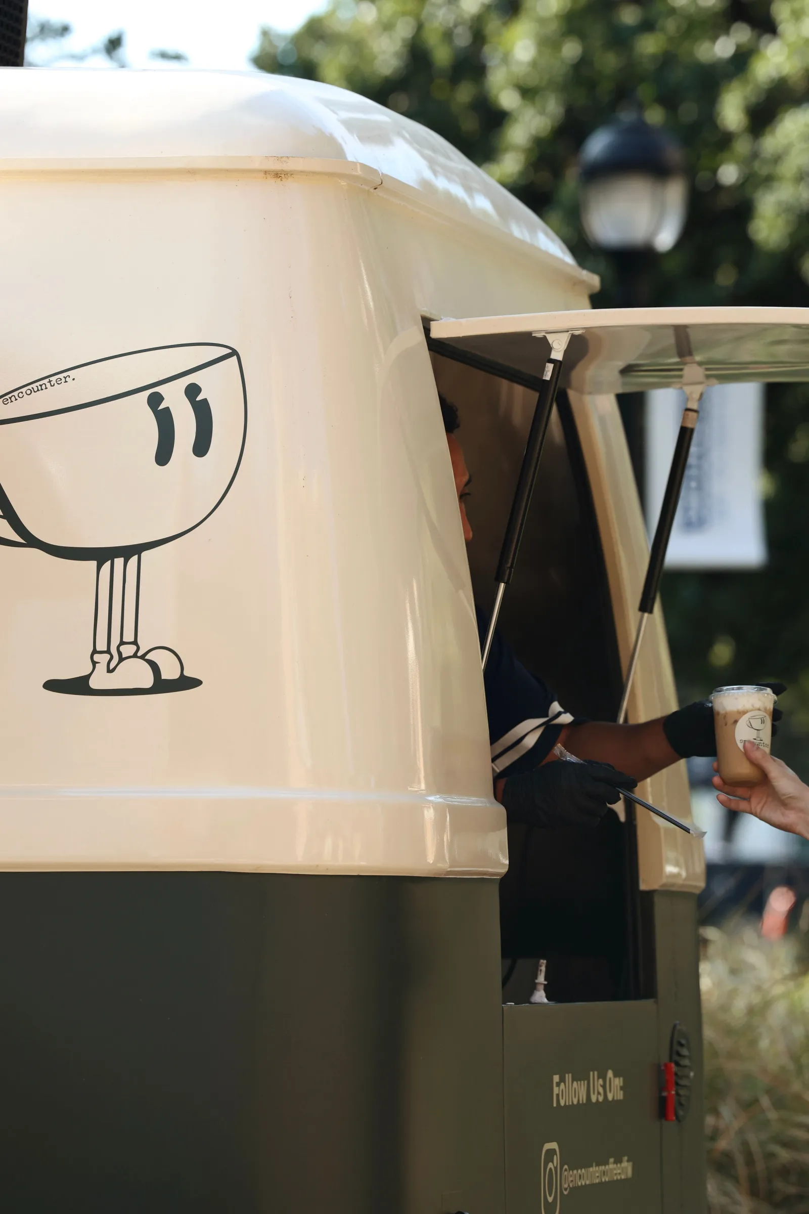 A barista handing an iced latte to a guest through the serving window of the Encounter Coffee VW bus, beside the coffee-cup mascot logo